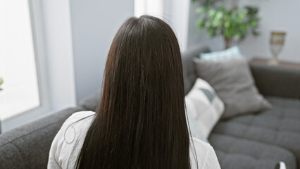Rearview of a young hispanic woman with long brunette hair, sitting in a cozy modern living room, portraying a casual, domestic lifestyle.