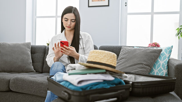A young woman checks her smartphone while packing a suitcase at home, indicating preparation for travel.