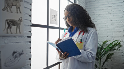 An african american female veterinarian in a clinic taking notes in a notebook with diagrams of animals on the wall.