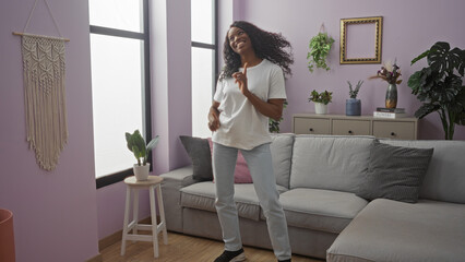 A beautiful african american woman with curly hair is happily dancing in her living room wearing a...