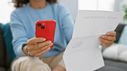 A mature woman reviews employment bills while holding a smartphone in her well-lit living room.