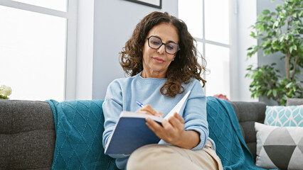 Mature woman with curly hair writing in a notebook while sitting on a blue sofa indoors.