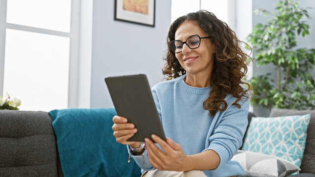 Smiling woman with curly hair wearing glasses and blue sweater using a tablet in a cozy living room