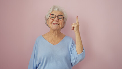 A cheerful senior woman with glasses pointing upwards against a pink isolated background.