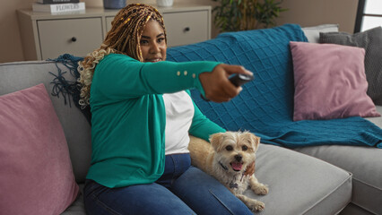 Young african american woman with braids sitting in a cozy living room, holding a remote and petting a dog.