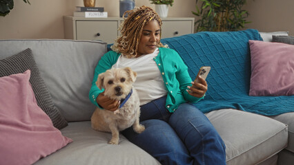 Young african american woman with braids sitting in a living room holding a small dog while looking at her smartphone indoors.