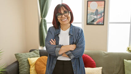 Obraz premium A confident middle-aged hispanic woman stands with arms crossed in a cozy living room, exuding warmth and comfort.