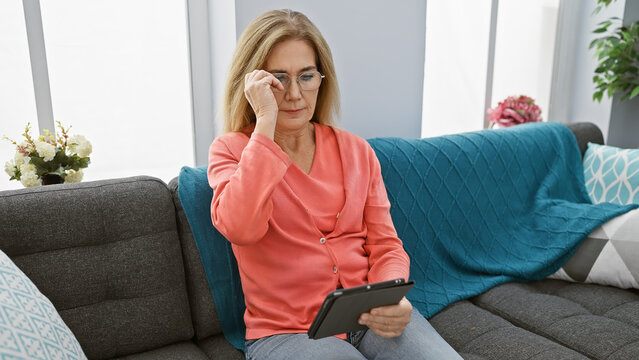 Mature blonde woman wearing glasses feeling stressed while holding a tablet in her cozy living room.