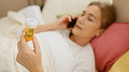 Fototapeta premium A middle-aged woman examines medication while talking on a phone in a bedroom setting.