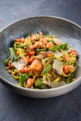Traditionelle italienische Pasta mit grünem Spargel, Garnelen und geräucherten Lachsstreifen serviert mit Pinienkernen und Parmesan Scheiben als Nahaufnahme in einer Nordic Design Schale
