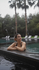 Fototapeta premium A contemplative young woman relaxes poolside at a luxurious bali resort, set against a backdrop of tropical palms.