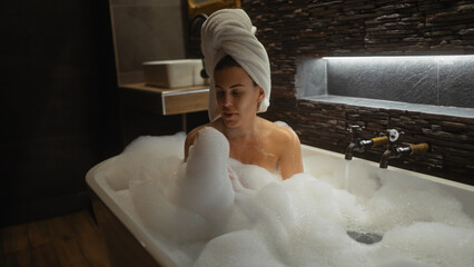 A young woman enjoys a relaxing bubble bath in a modern bathroom with a towel on her head.