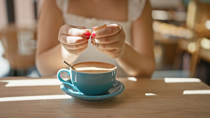 Caucasian young woman captivated by the aroma of her morning espresso, pouring sugar into her coffee cup with hands, sweetening the hot beverage at a sunny cafe table, inside the cafeteria.