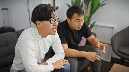 Two asian men engaged in an animated discussion while sitting in a modern living room with a couch and plants in the background, depicting friendship or brotherhood in an indoor setting.
