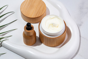 Bamboo cream jar and dropper bottle with face oil on a tray on marble background. Cosmetic container mockup. Background for branding and packaging presentation. Natural skincare beauty product concept