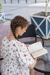 Young caucasian girl in a casual dress, reading book in street cafe