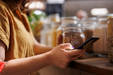 Green living woman on a diet using smartphone to make sure zero waste supermarket food is good for her health, close up. Client in local sustainable store making sure products are organic
