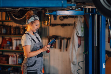 Female mechanic with tablet under lifted car doing car diagnostics.