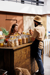 Client shopping for organic pantry staples in zero waste store using plastic free paper bag, being assisted with information by trader. Seller showing woman products with no artificial ingredients