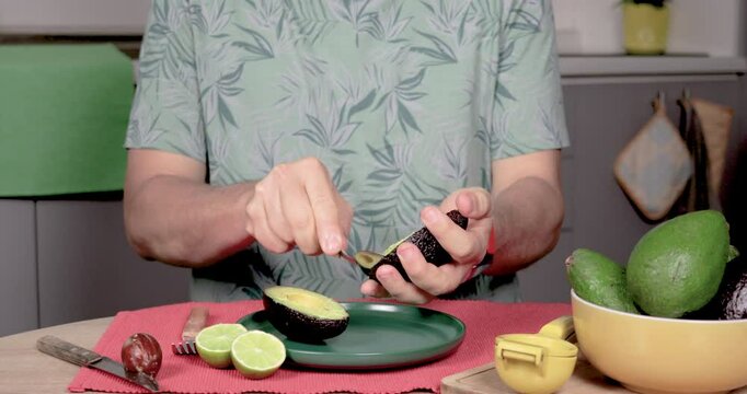 4K video of a man sitting at a table, scooping a Hass palta with a spoon and placing it in a plate. Predominantly green, with red, yellow, and gray. Concept of homemade preparation
