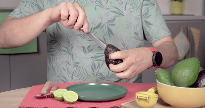 4K video of a man sitting at a table, opening a Hass palta with a knife and removing the pit. Predominantly green, with red, yellow, and gray. Concept of homemade preparation