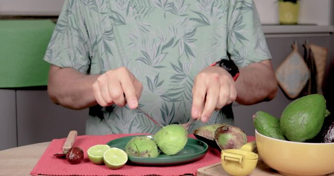 4K video of a man sitting at a table, slicing and mashing a Hass palta with a fork in a plate. Predominantly green, with red, yellow, and gray. Concept of homemade preparation