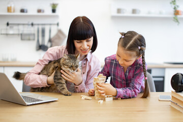 Cat sitting on mothers lap, laptop and smartphone on table. Bright kitchen background. Mother and daughter enjoying quality time together at home, playing a game with wooden blocks on table.