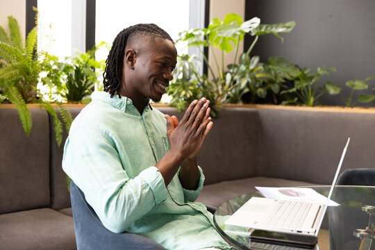 Smiling man using laptop and video conferencing from home office, hands clasped