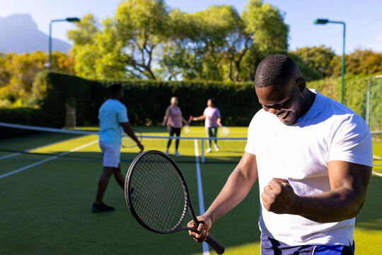 Celebrating victory, man holding tennis racket with diverse friends playing in background