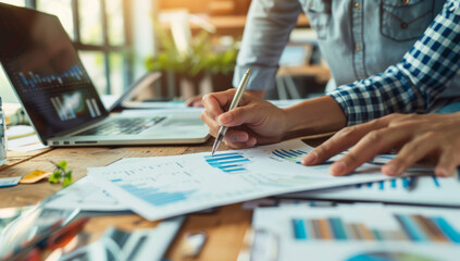 A person is pointing at the document on their desk while another individual holds out a pen to sign it.