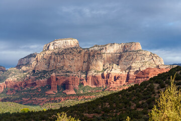 A majestic red rock mountain formation in Sedona, Arizona, with dramatic lighting from a partly cloudy sky.