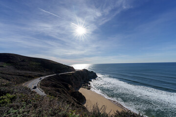 A winding coastal highway hugs a rugged California coastline, overlooking a sandy beach with crashing waves under a bright sun.