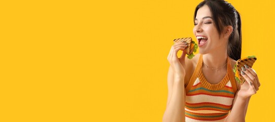 Happy young woman eating tasty sandwiches on yellow background with space for text