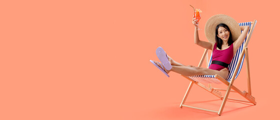 Beautiful young Asian woman in swimsuit with tasty aperol spritz sitting on deckchair against orange background