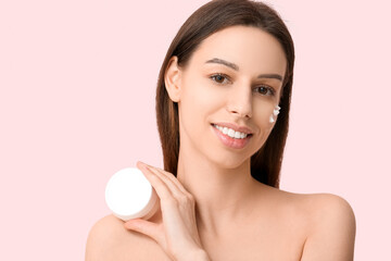 Beautiful young woman with cream on cheek holding jar of sunscreen against pink background