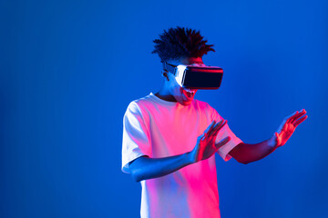 Young African American gaming player wearing VR pointing hologram 3D graphic isolated blue pink...
