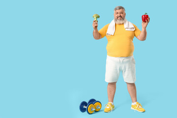 Overweight happy mature man with vegetables and dumbbells on blue background. Weight loss concept