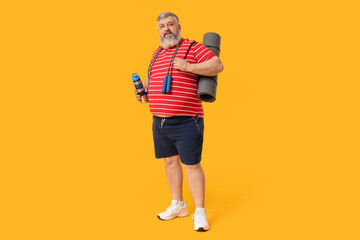 Fototapeta premium Overweight mature man with jumping rope, yoga mat and bottle of water on yellow background. Weight loss concept