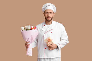 Young male chef with flowers and gift box on beige background. International Women's Day