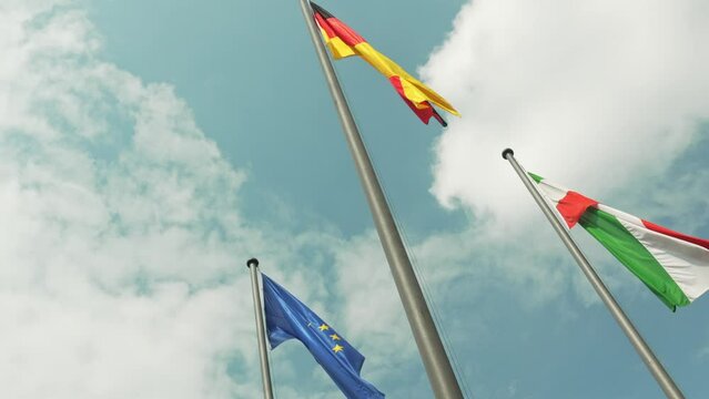The flags of Germany, Italy and the European Union on flagpoles flutter in the wind against the background of the sky and lands, slow motion