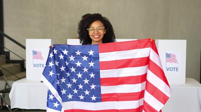 African American woman smiles waving the United States american flag at a US election polling station. In the background voting booths place, democracy in America