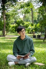 A nerdy young Asian male college student wearing eyeglasses is sitting on the grass in the campus park with a book.
