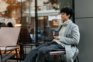 Stylish young man sitting on a chair with a coffee cup in one hand and a phone in the other, in front of a building