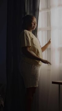 Vertical full shot of young Black woman looking out of window at dawn and walking around room on sleepless night. Avoiding real life, feeling lonely, living with ptsd