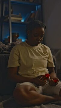 Vertical full shot of African American lady in homewear sitting on floor by unmade bed and hesitating to take tranquilizers at dawn after sleepless night