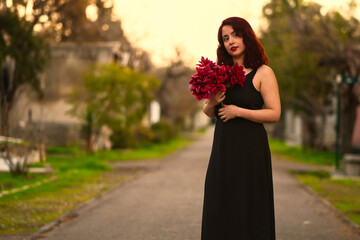 Fototapeta premium Beautiful woman with long black dress, holding bouquet of red flowers, in a cemetery, sad and melancholic look. in an outdoor forest. Halloween party and celebration.