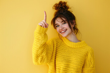 Girl with raised index finger wearing yellow sweater on yellow background. Pointing gesture on yellow background. Smiling woman with raised finger upwards. Attention. 
