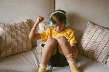 Little boy enjoying his free time using digital tablet has happy emotion and wears yellow t-shirt using green headphones.