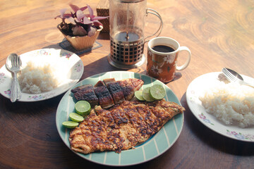 Overhead outdoor breakfast spread of grilled fish, omelette, rice, lime wedges and coffee in a French press 
