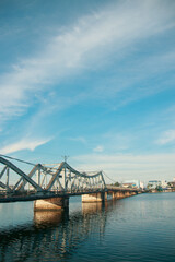 A calm river flows under a the famous old bridge in Kampot town in Cambodia, reflecting the clear blue sky and scattered clouds, with a cityscape in the background